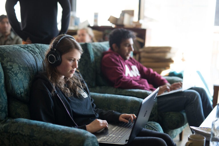 A young white woman sits on her laptop in a cafe, wearing headphones.