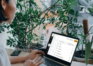 A woman works on a laptop in a cafe with lots of plants around.